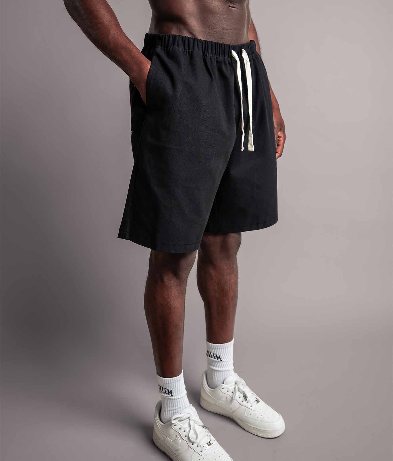 Schräge Perspektive vom Model das die schwarzen TELLEM Baumwoll Shorts aus Cotton Twill mit lockerem Fit trägt und Hand in Hosentasche hat.
